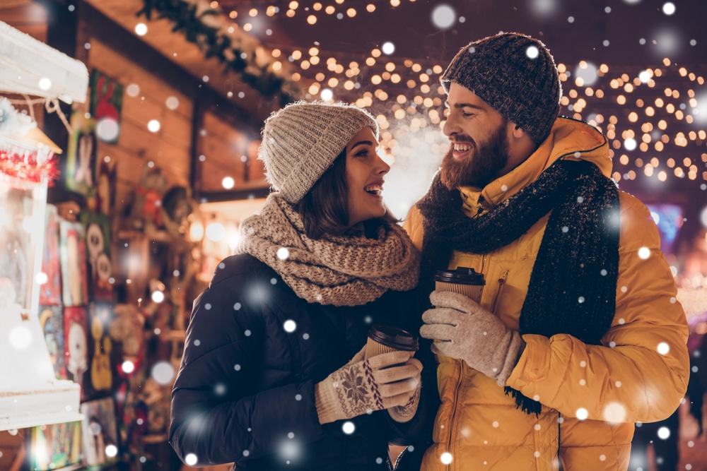 Woman and man, all bundled up in winter outerwear walk through downtown lights holding hot cocoa on their Michigan Winter Getaways for Couples