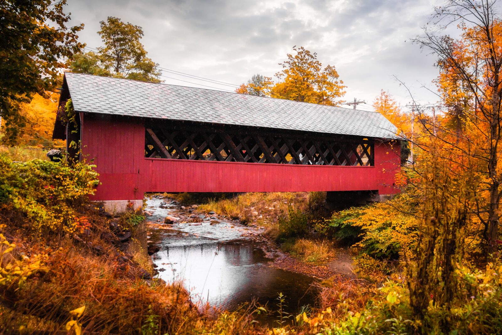 A red covered bridge over small river, surrounded by fall foliage in Vermont.
