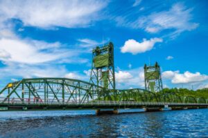 A green vertical-lift truss bridge stands over a blue river against a bright blue sky with white clouds.
