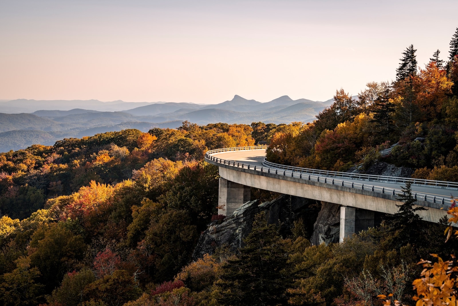 A viaduct bridge hugging a mountain of fall colors in North Carolina with blue mountains in the distance on the Blue Ridge Parkway.