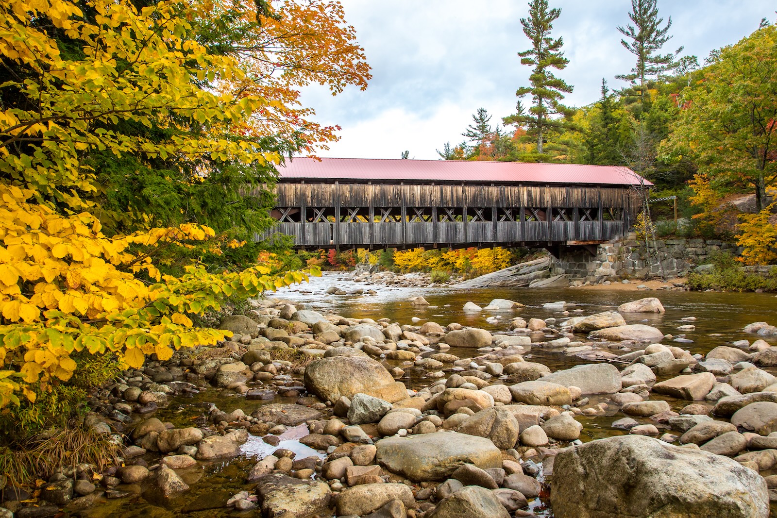 The Albany Covered Bridge, one of the best covered bridges in New Hampshire, surrounded by fall foliage and a rocky river.
