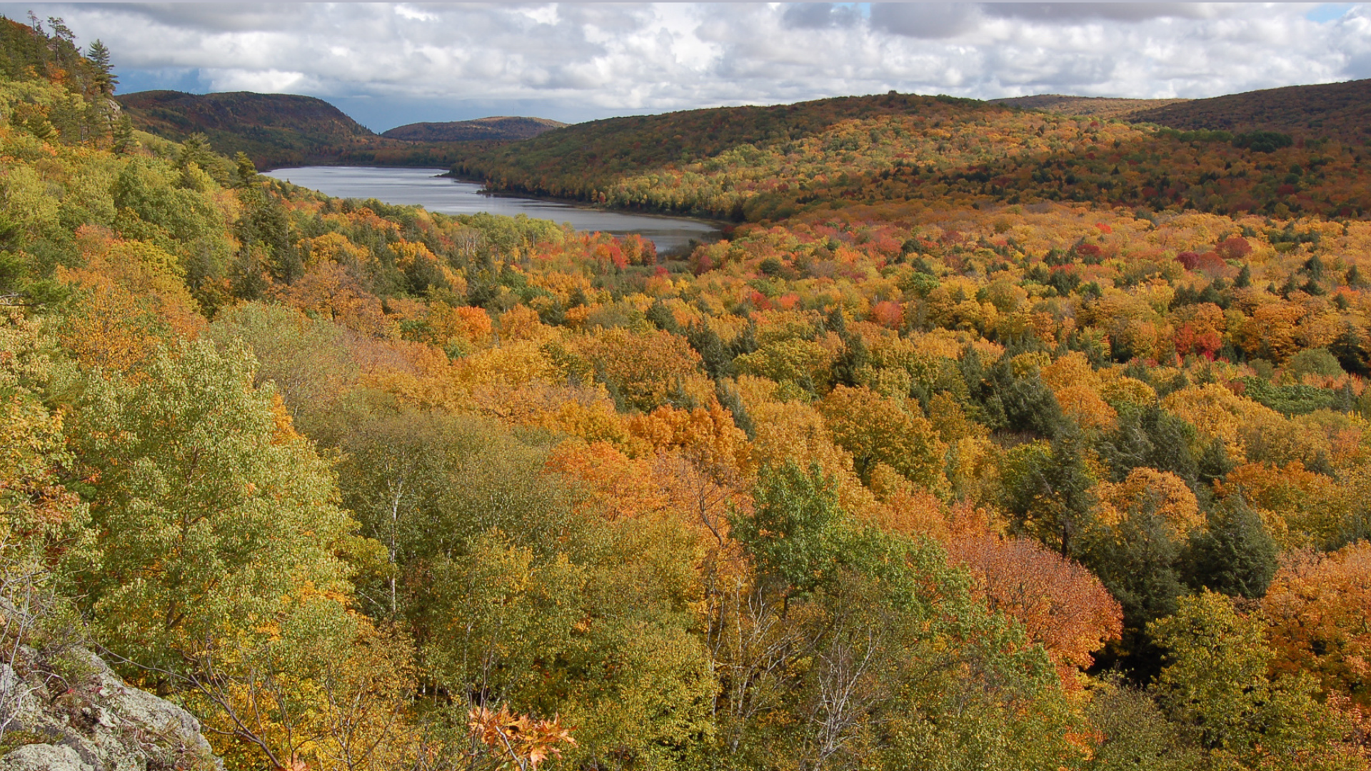 Panoramic view of a lake surrounded by rolling hills and a forest ablaze with vibrant autumn colors.