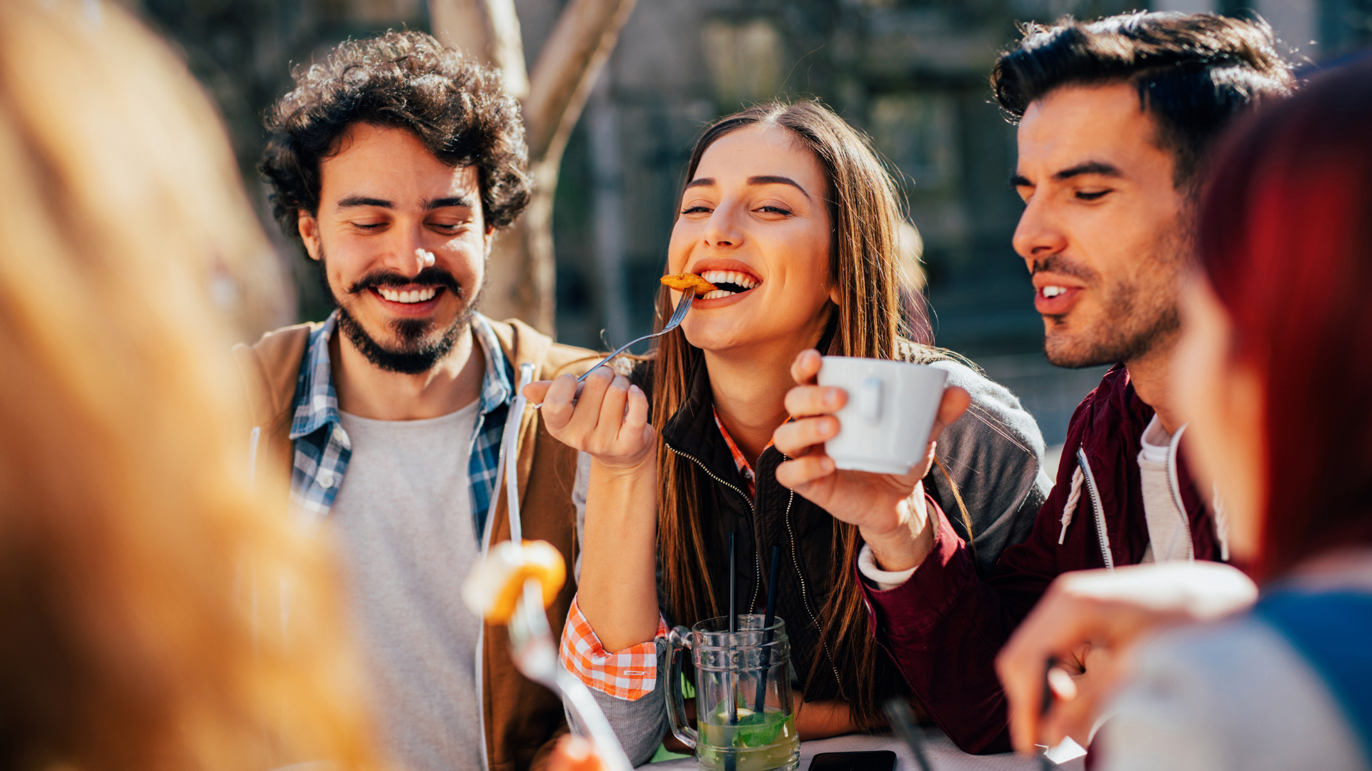 Friends enjoying a casual outdoor meal together, laughing and sharing food and drinks around a table.