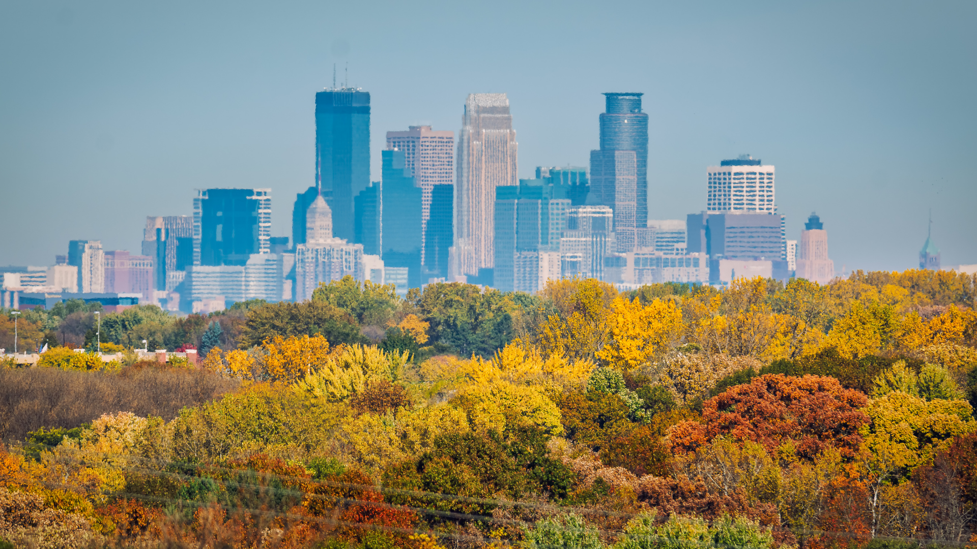 Minneapolis skyline rising behind a colorful autumn landscape of trees and foliage.