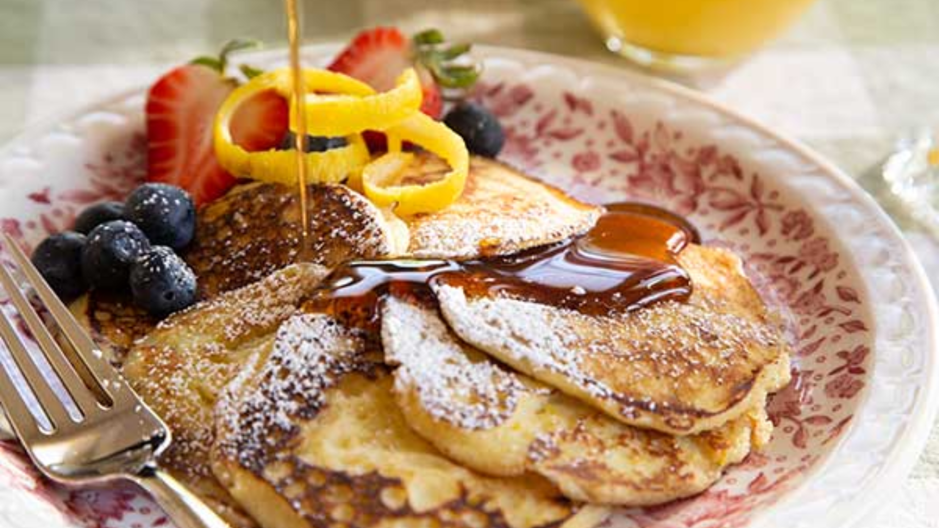 A breakfast plate of pancakes topped with fruits, syrup, and powdered sugar.