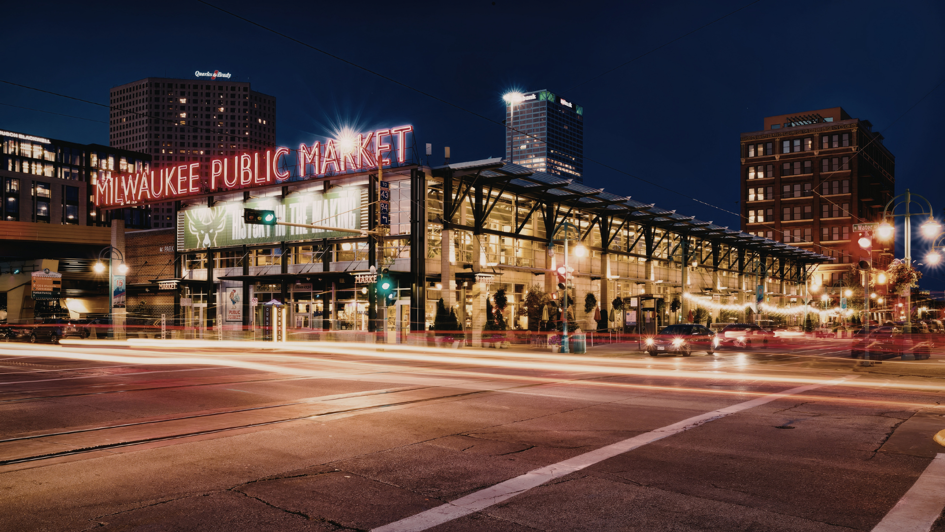 Milwaukee Public Market illuminated at night, with downtown buildings and light trails from passing traffic in the foreground.