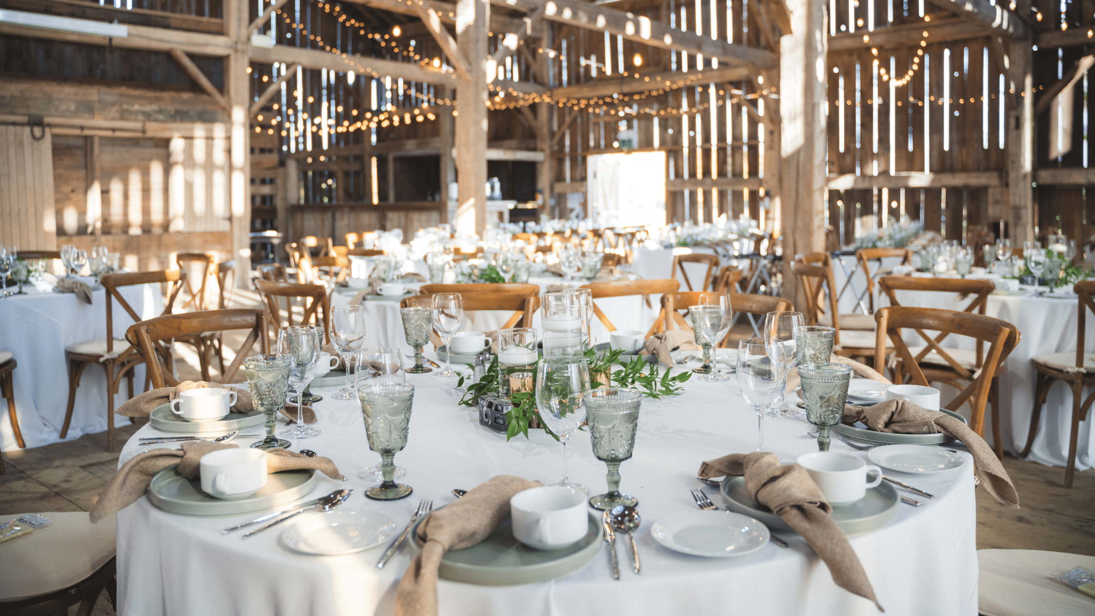 A beautifully decorated wedding reception in a rustic wooden barn, with round tables covered in white linens and set with white plates, wine glasses, textured goblets, folded tan napkins, and greenery centerpieces. Warm string lights hang throughout the barn, creating a bright, elegant atmosphere.