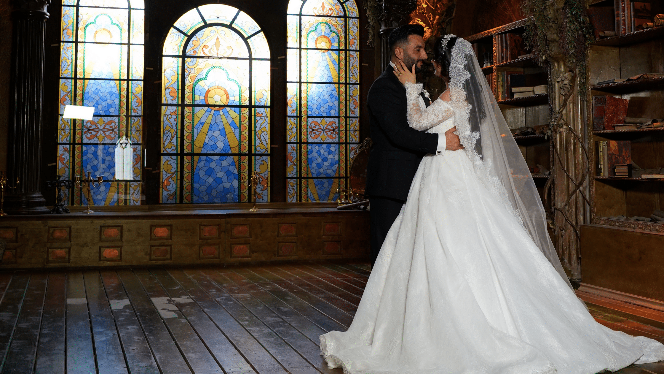 Newlywed couple embraces in a historic library, with the bride in a flowing white gown before colorful stained-glass windows.
