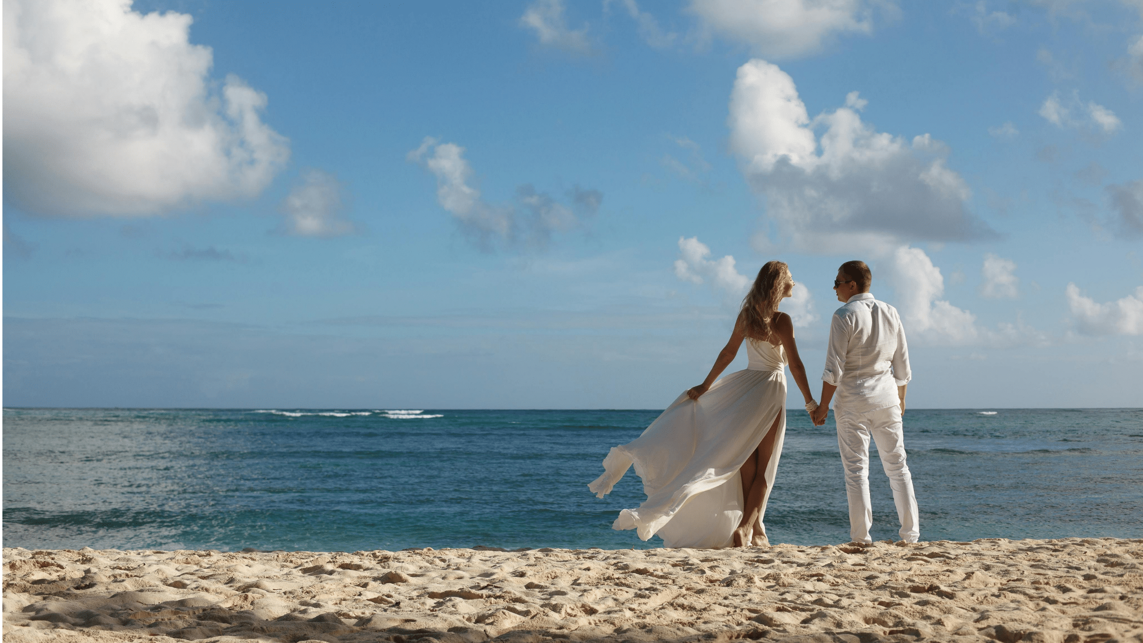 A newlywed couple in wedding attire walks hand in hand along a sandy beach beside the ocean, with the bride’s white dress flowing in the breeze. A bright blue sky with scattered clouds stretches above the calm water.