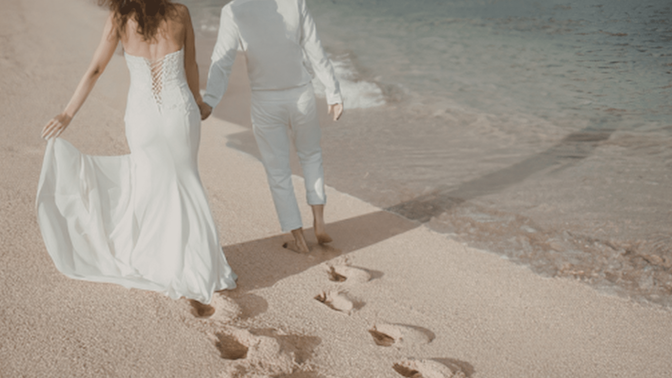 Newlywed couple in white wedding attire walks hand in hand along a sandy beach, leaving footprints by the gentle waves in a serene scene.