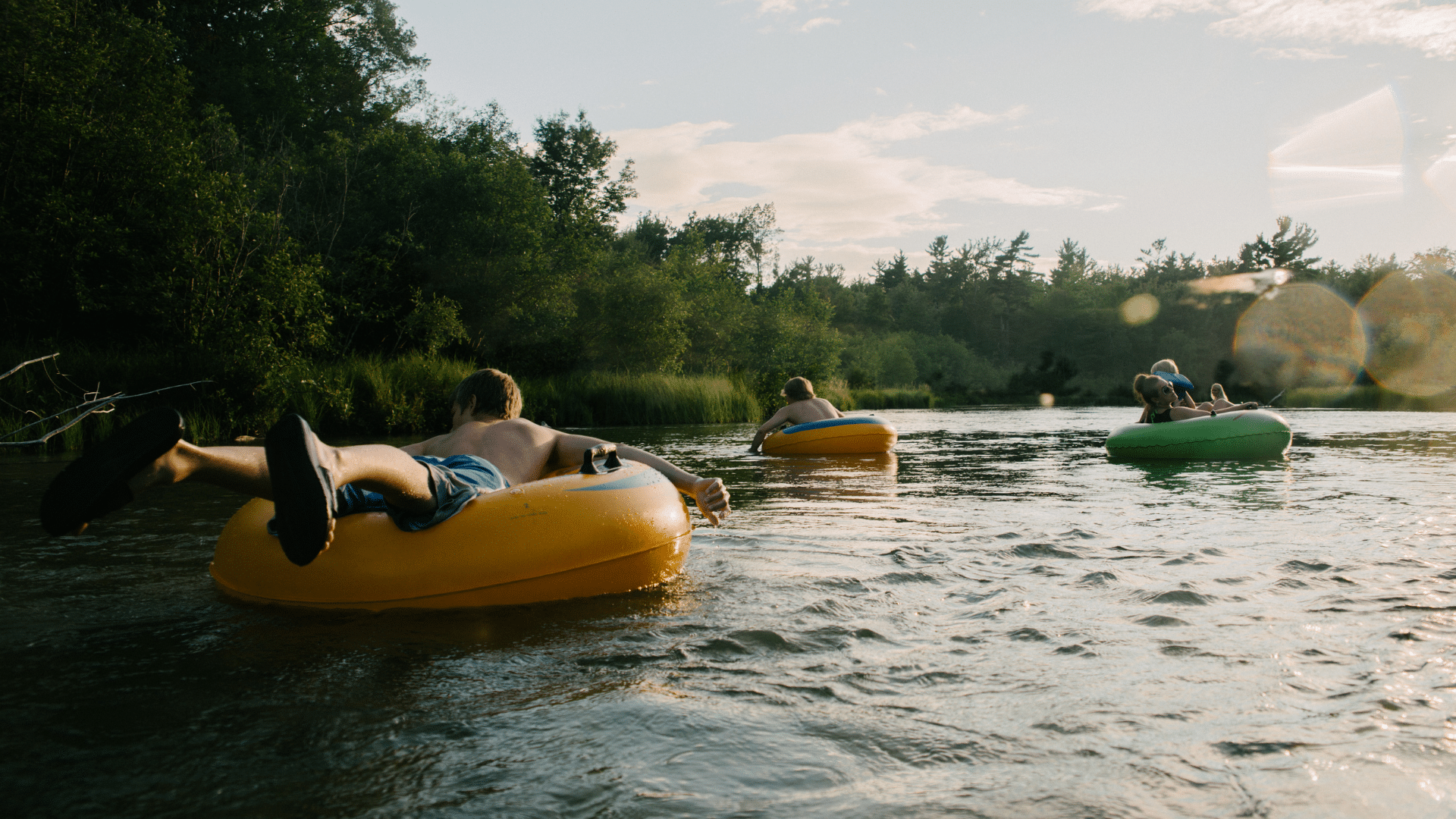 Three people relax on inflatable tubes while floating down a calm, tree-lined river on a sunny summer afternoon. A person in a yellow tube is in the foreground, with two others farther downstream in yellow and green tubes.