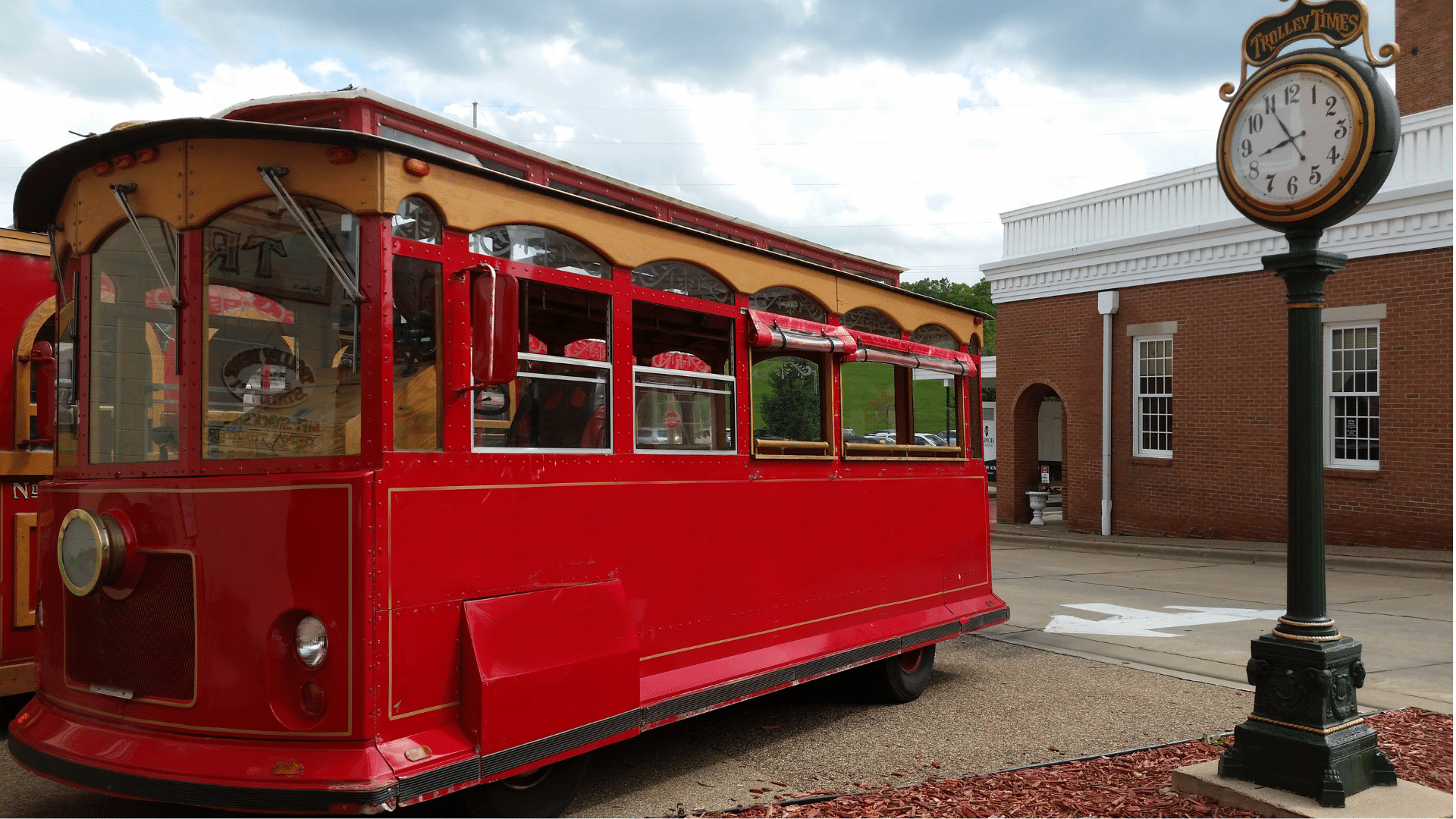 A bright red vintage streetcar with wood trim is parked along a downtown street in Galena, Illinois. A decorative trolley-themed clock stands beside a historic red-brick building, with cloudy skies and green hills visible in the background.