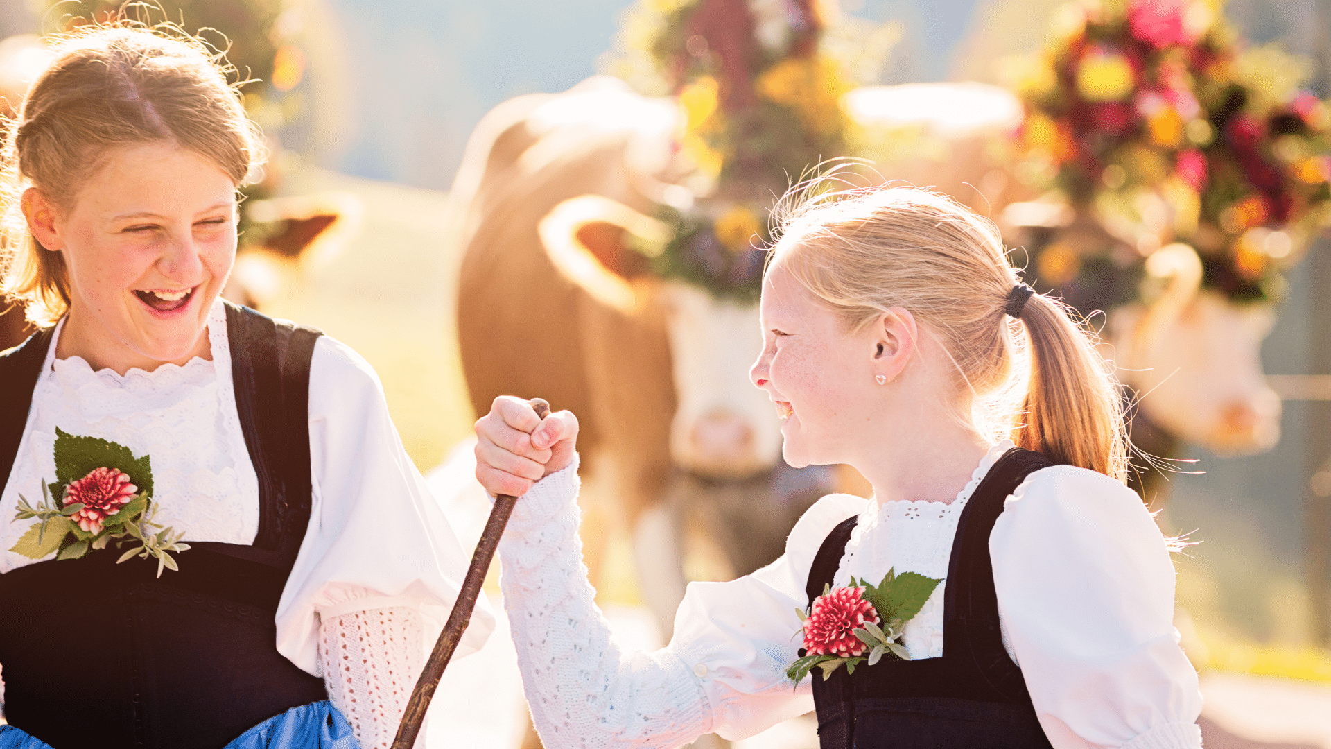Two young girls in traditional folk clothing laugh together outdoors at a sunny festival, each wearing a floral decoration on her outfit; one holds a wooden walking stick.