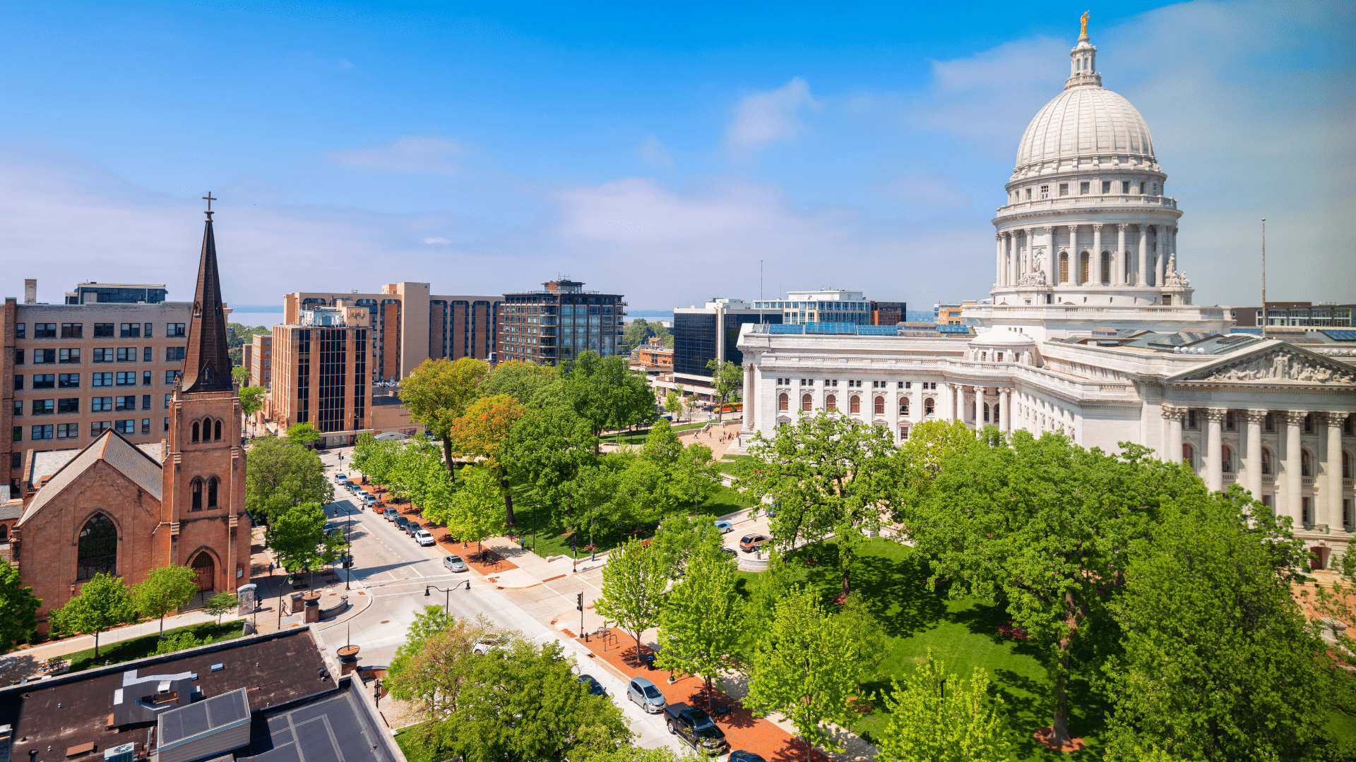 Sunny aerial view of downtown Madison, Wisconsin, with the State Capitol dome, historic church, tree-lined streets, and colorful city buildings.