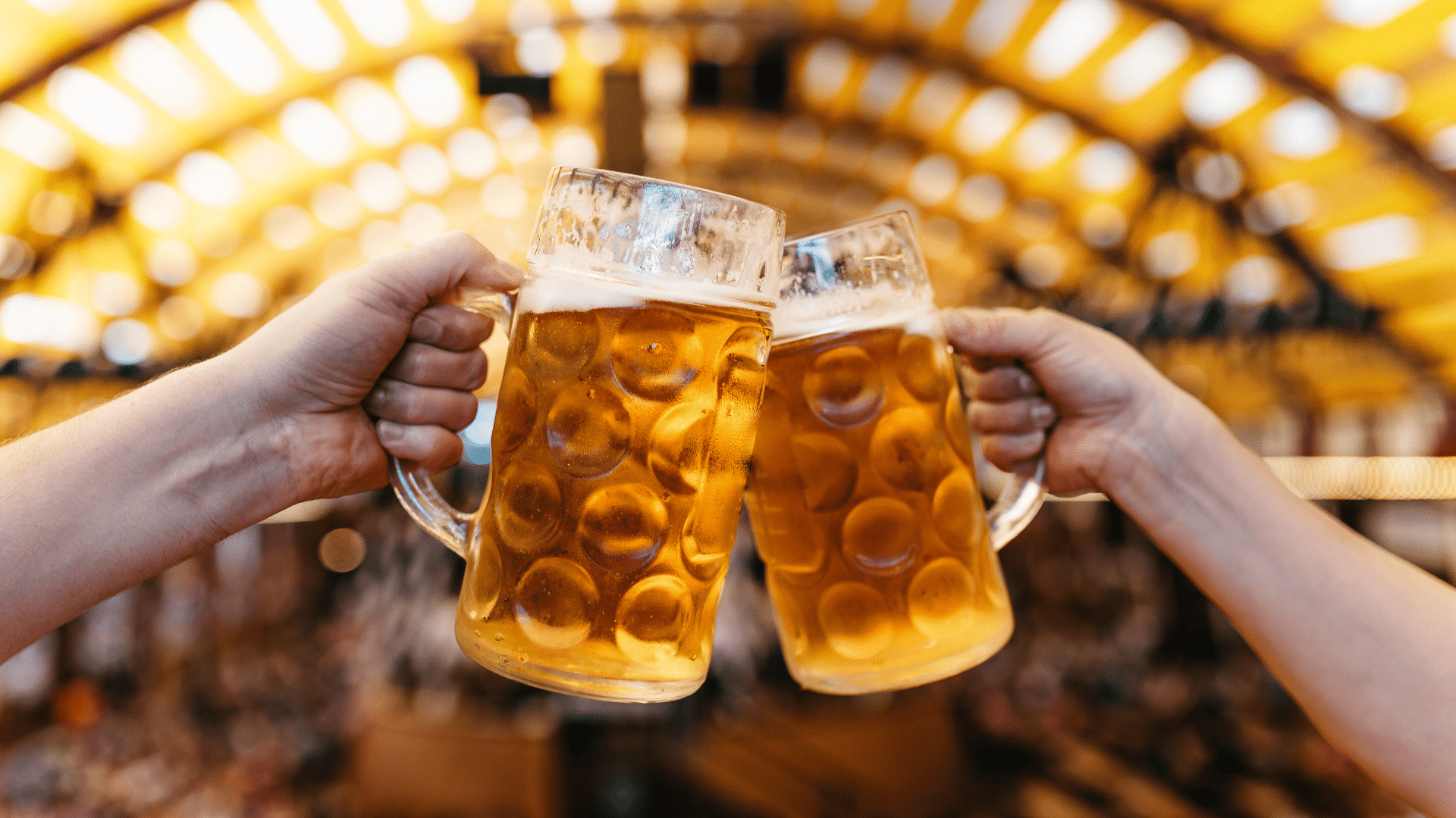 Two hands raise frosty mugs of golden beer in a toast at a lively Oktoberfest celebration, with warm golden lights and a festive crowd in the background.