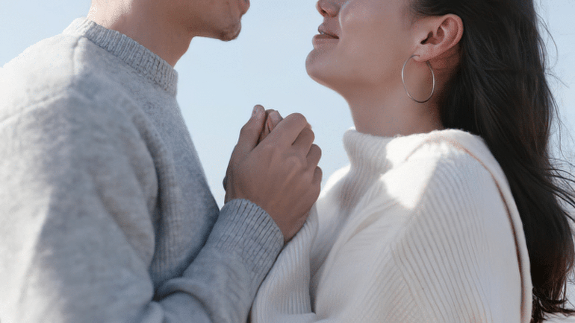 Close-up of a couple in light-colored sweaters holding hands and facing each other affectionately outdoors, with a soft blue sky in the background, emulating romantic midwest getaways.