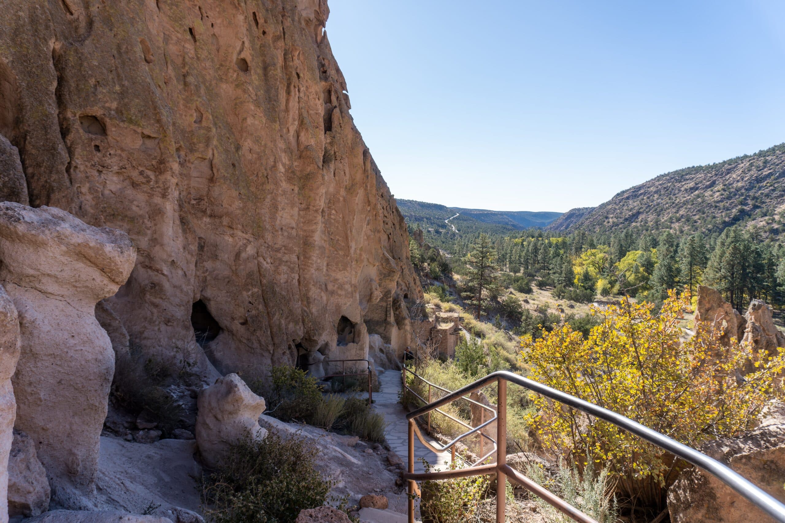 The Bandelier cliff dwellings at the Bandelier National Monument with a walking path and rail beside the dwellings and foliage.