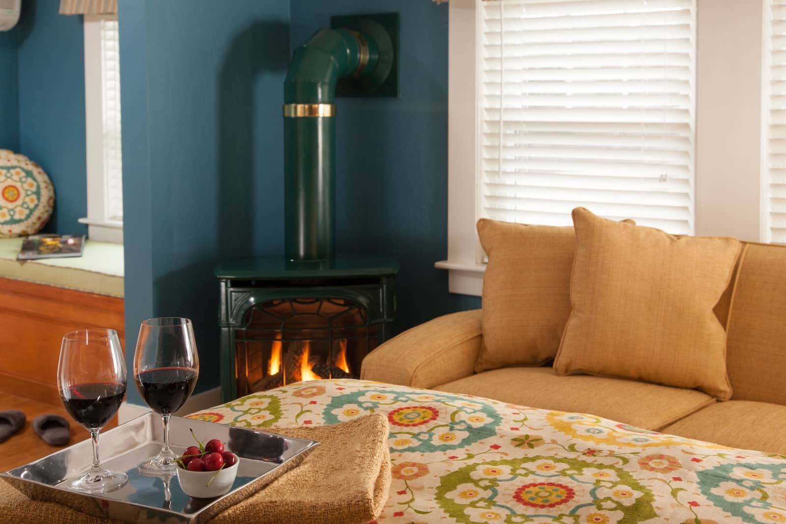 Two glasses of wine on the edge of a bed in front of a couch and a fire place in a guest room at a New Hampshire Bed and Breakfast.