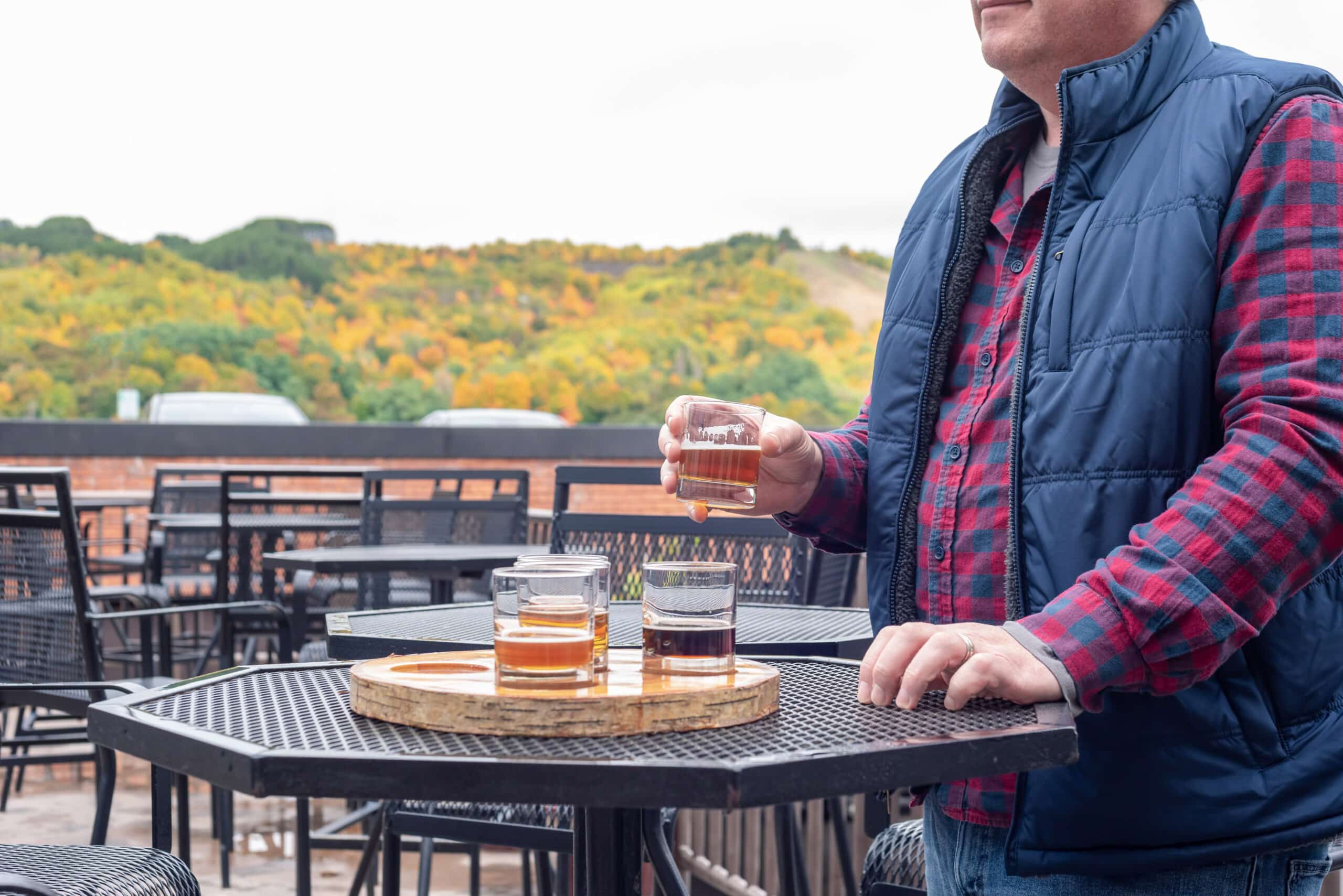 Person enjoying a craft beer tasting flight on an outdoor patio, with several beer samples on a wooden serving board and colorful autumn foliage in the background.