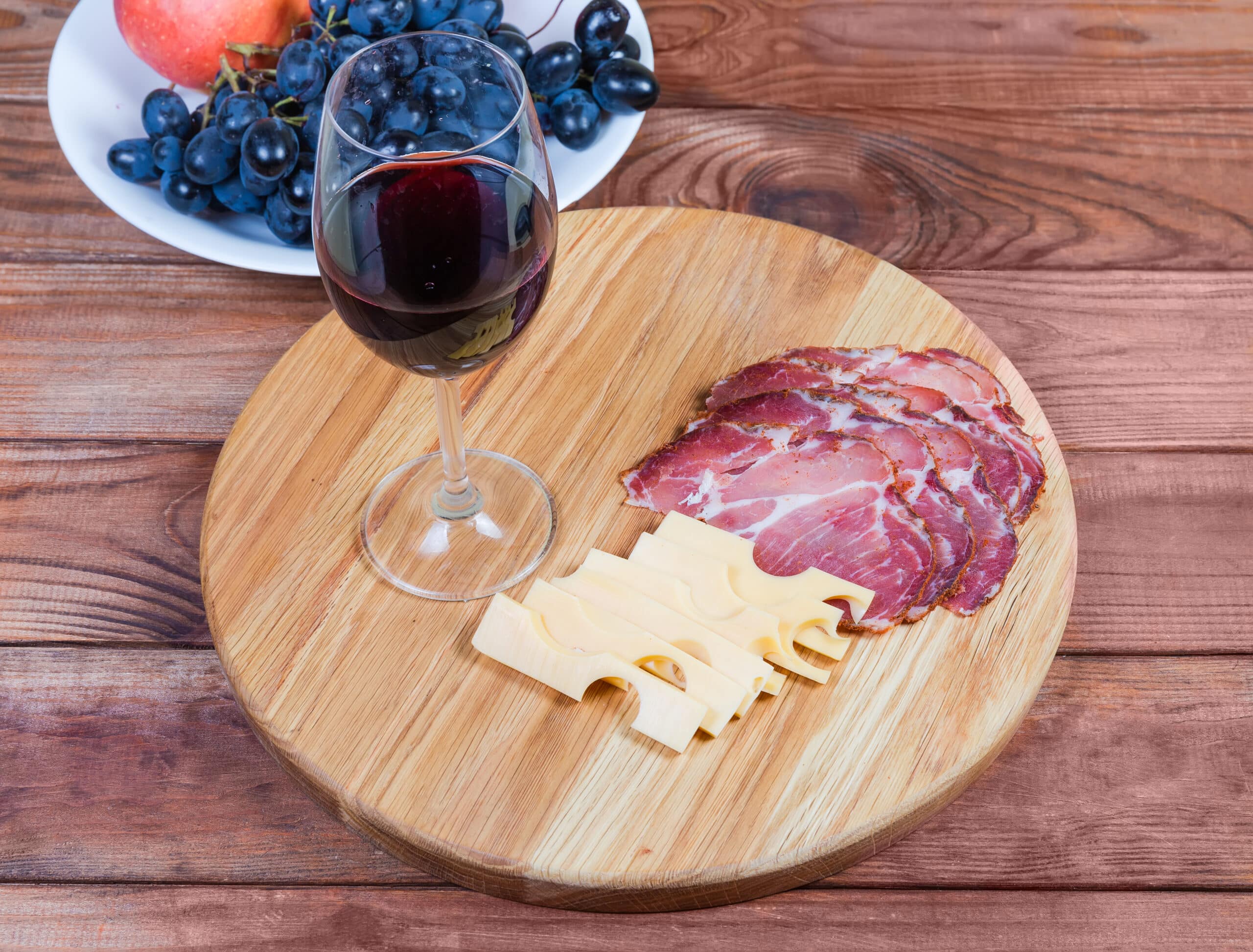 A glass of red wine, sliced cured meat, and pale cheese sit on a round wooden board beside grapes and an apple, creating a rustic, inviting snack spread.
