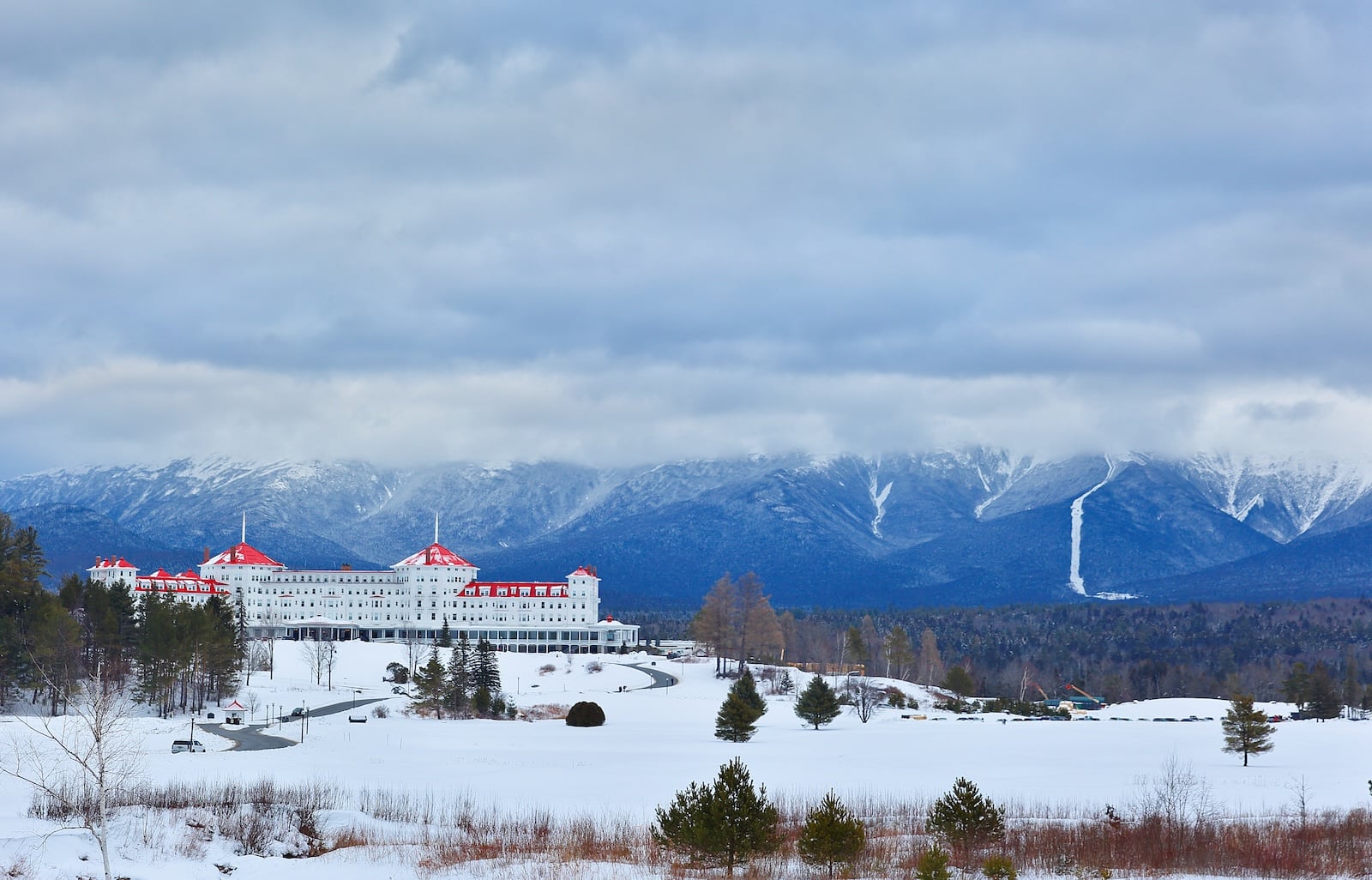 A white building with a red roof among snow and huge mountains, where you can find the Bretton Woods Nordic Center.
